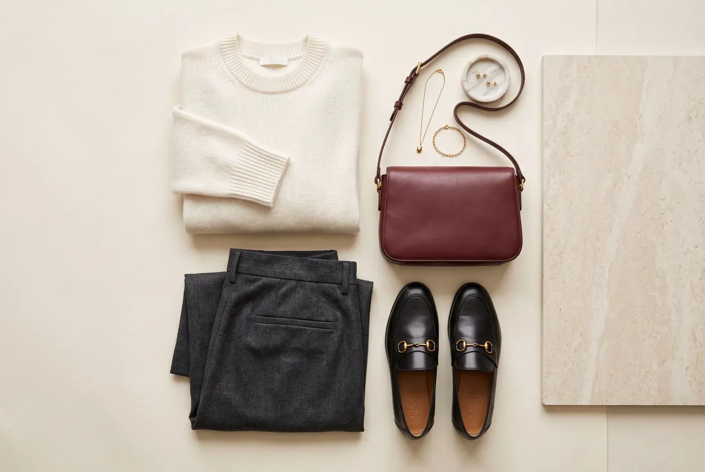 Editorial arrangement of a cream knit, tailored trousers, burgundy bag and black loafers